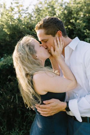 A romantic outdoor portrait of a couple sharing an intimate embrace and kiss in natural sunlight with greenery behind them.