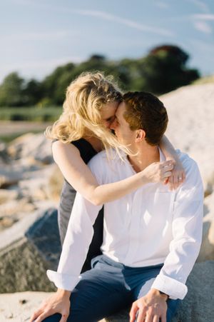 A romantic moment captured between a couple sharing a tender kiss on rocky terrain near the water on a sunny day.