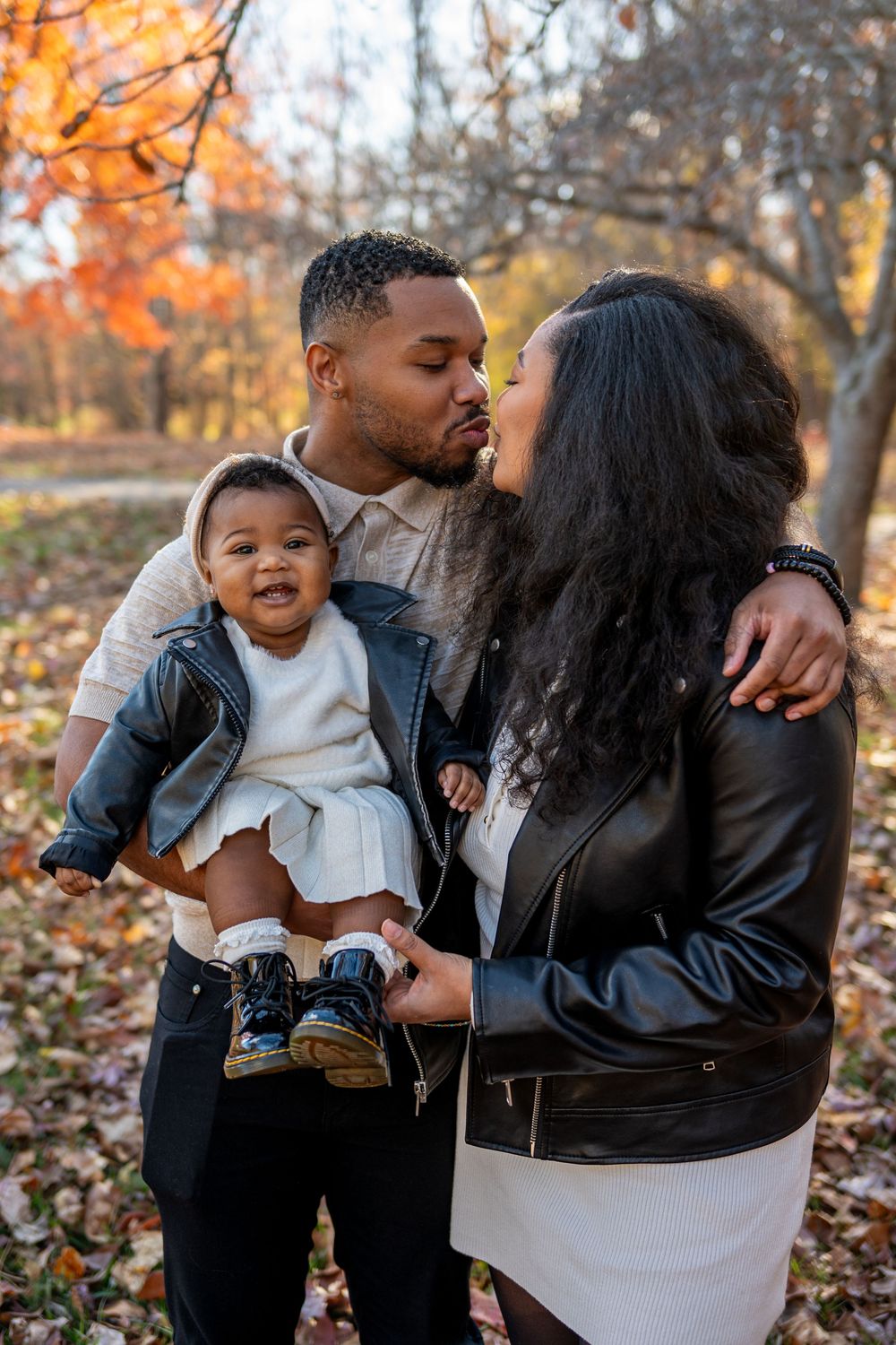 A happy family with a baby enjoys an autumn park, parents sharing a kiss while holding their smiling infant.