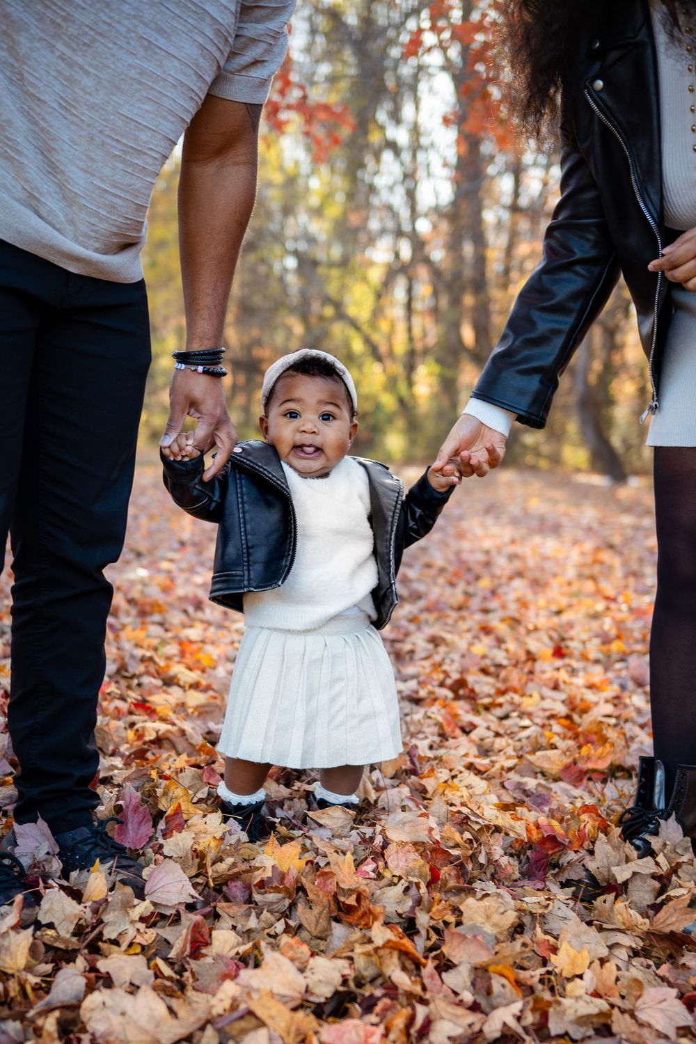 Toddler girl in white dress and leather jacket holds parents hands while standing in autumn leaves in a forest.