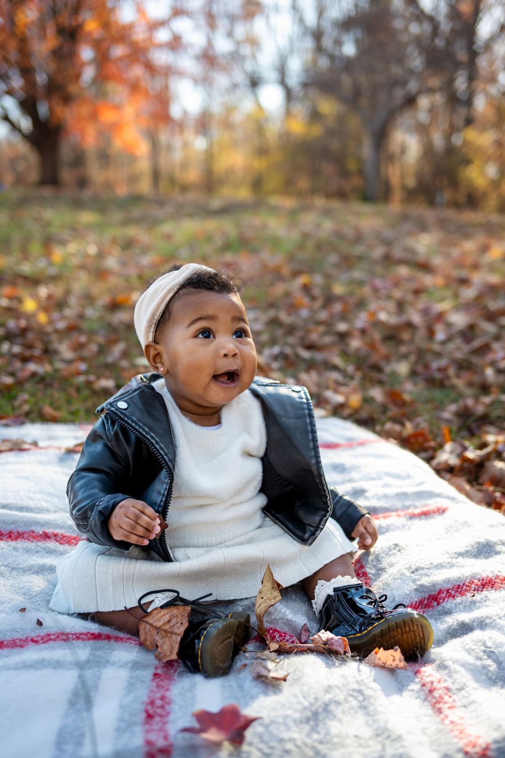Adorable baby in leather jacket and white dress sits on blanket among autumn leaves in a park.