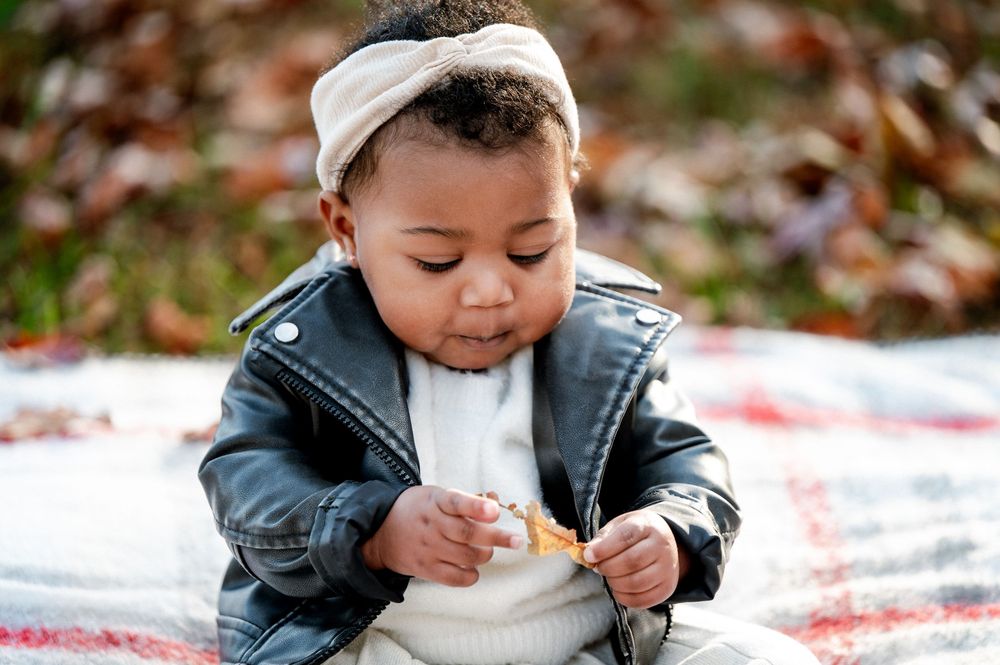 Adorable baby in leather jacket and headband sitting on blanket exploring a leaf outdoors.