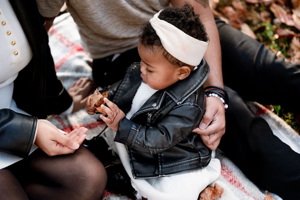 Adorable toddler in leather jacket and white headband sits outdoors during a family picnic in autumn.