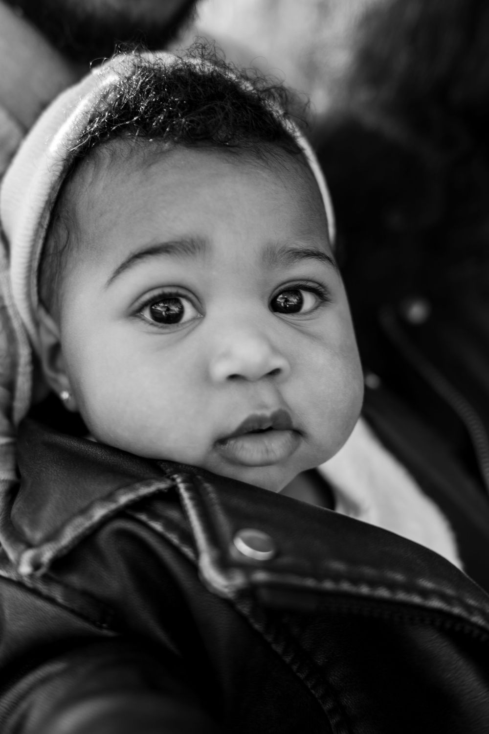 Adorable Black baby wearing a leather jacket and headband with big expressive eyes in a black and white portrait.