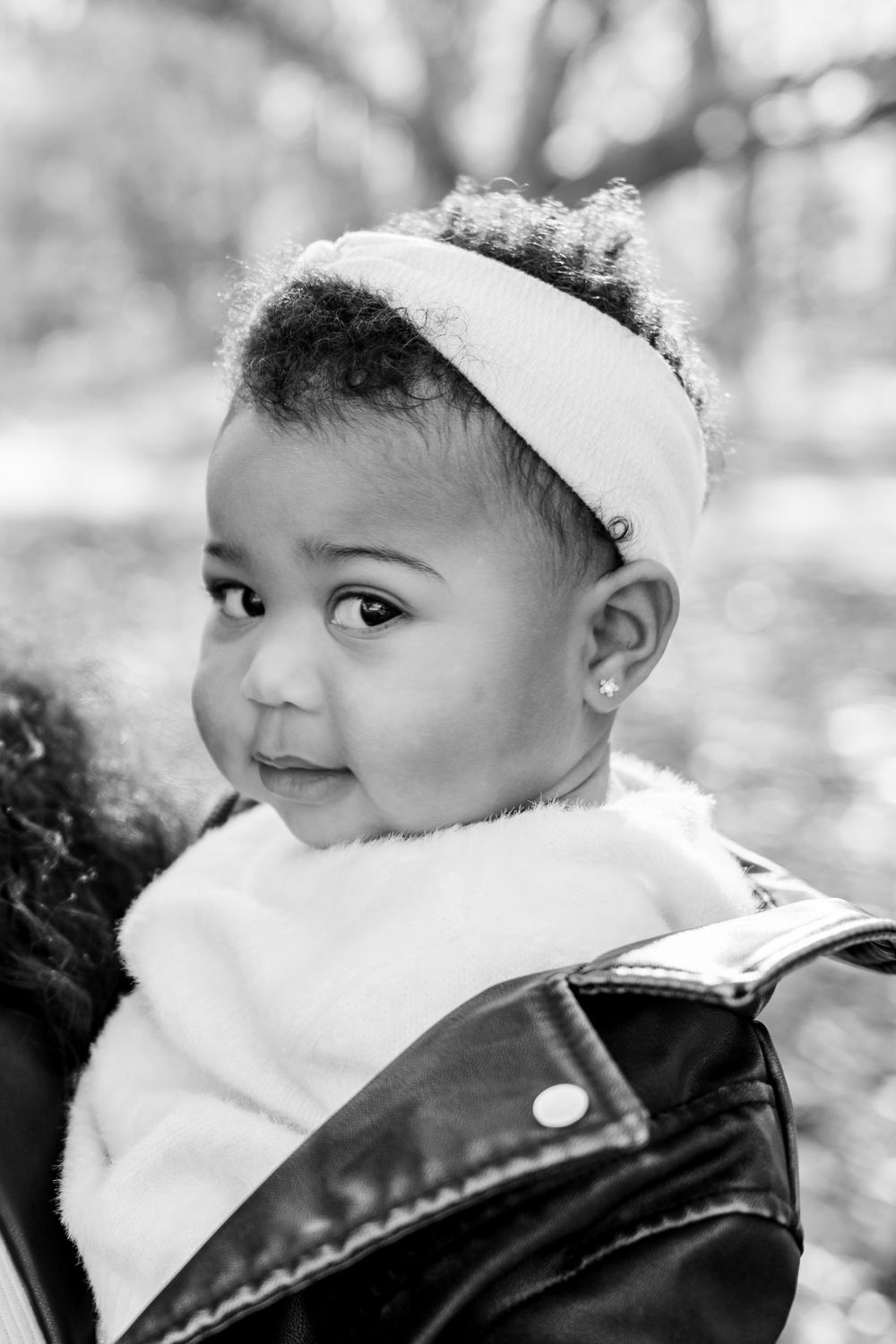 Black and white portrait of a cute baby girl wearing a headband and leather jacket, looking over her shoulder outdoors.