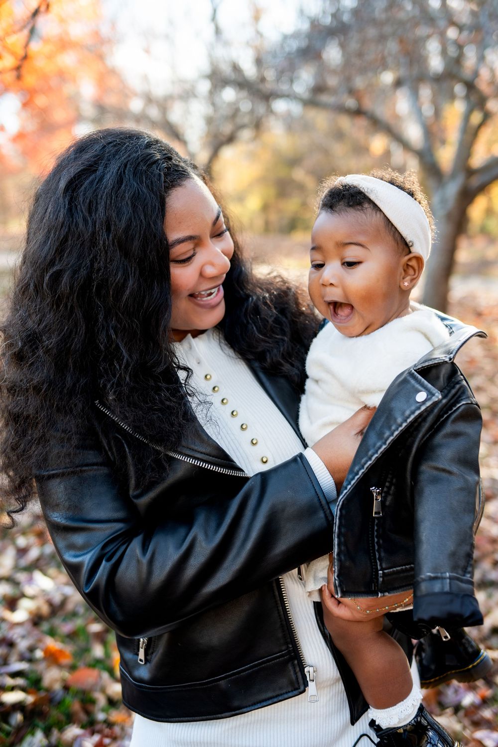 Joyful Black mother holding her laughing baby outdoors during autumn, both wearing matching leather jackets.