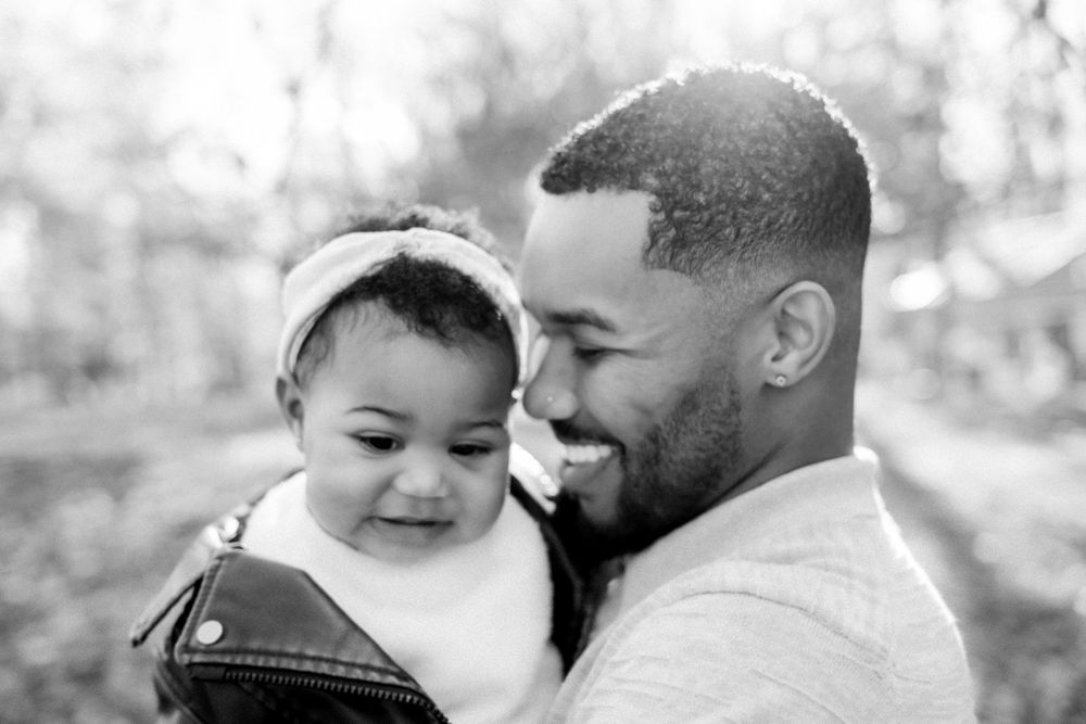 Black and white photo of a smiling father holding his baby girl outdoors among autumn trees.
