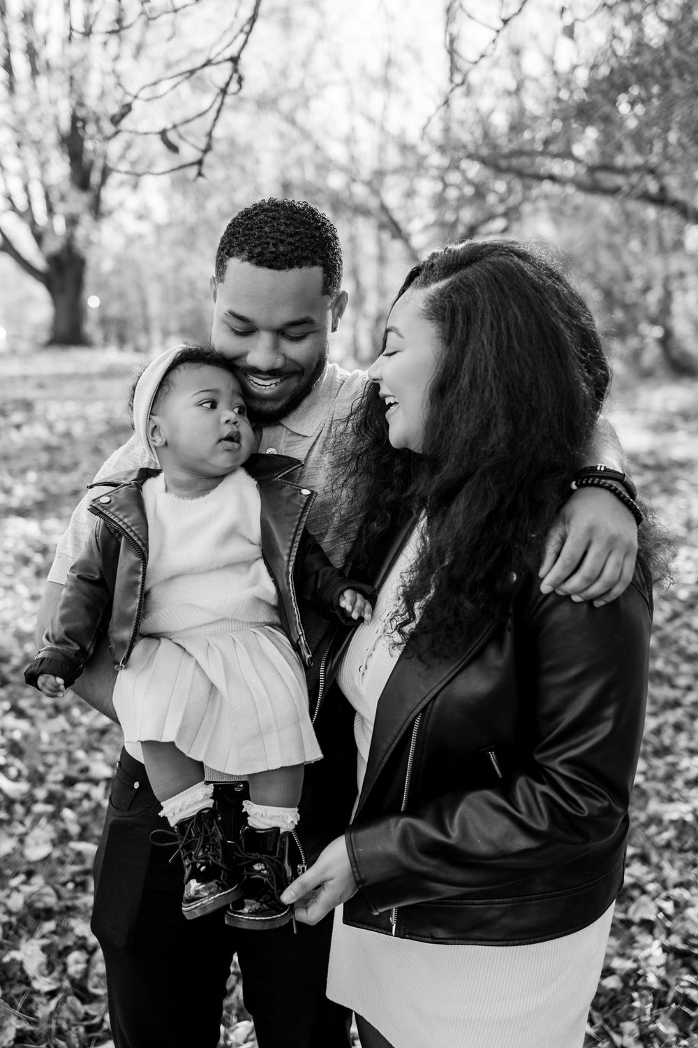 Black and white photo of a smiling family with parents holding their baby girl outdoors in a park during autumn.