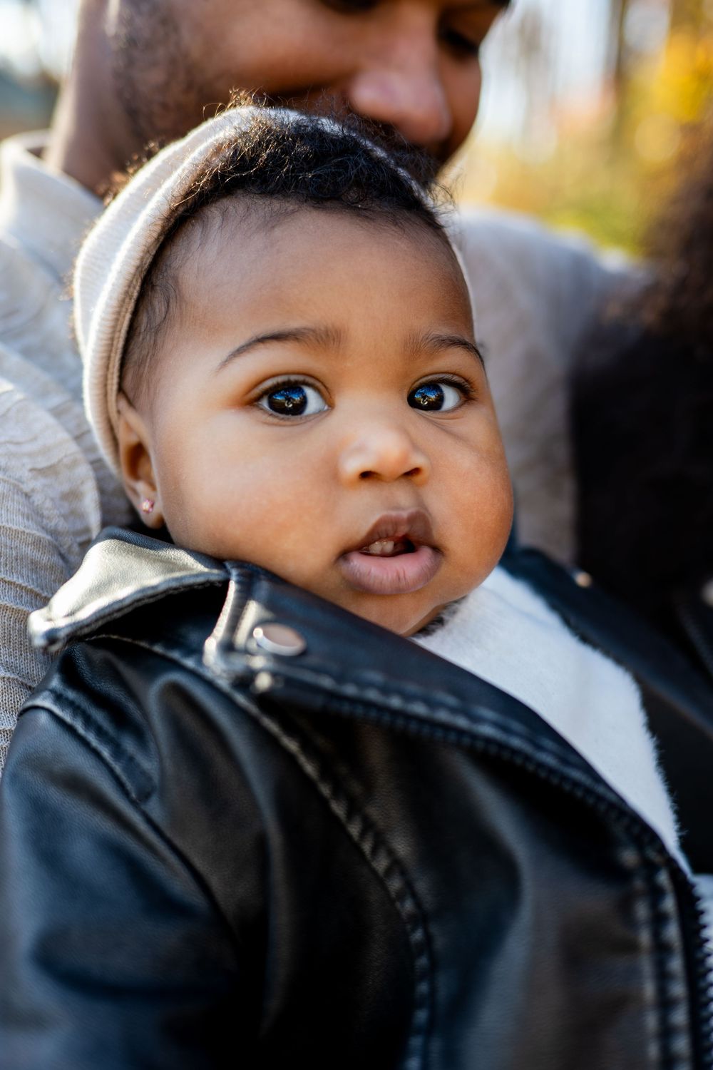Adorable Black baby in a leather jacket and beanie hat being held by a parent outdoors in autumn.