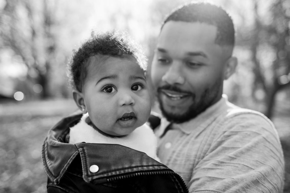 Black and white photo of a smiling father holding his curious baby outdoors in a park setting.