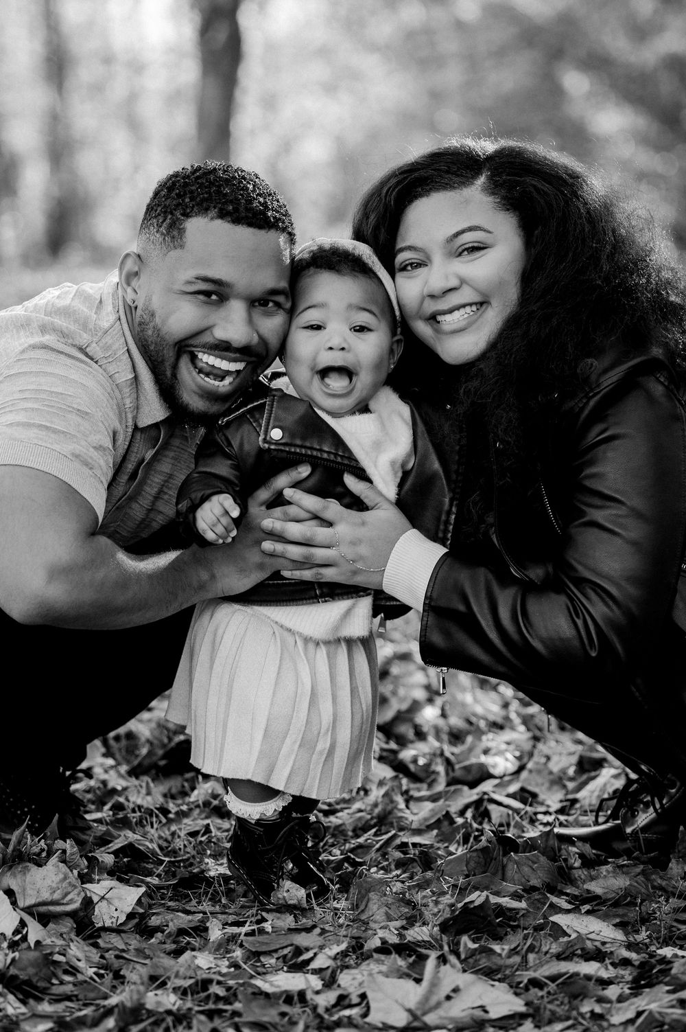 Happy Black family with mother, father, and baby laughing together among fall leaves in black and white photo.