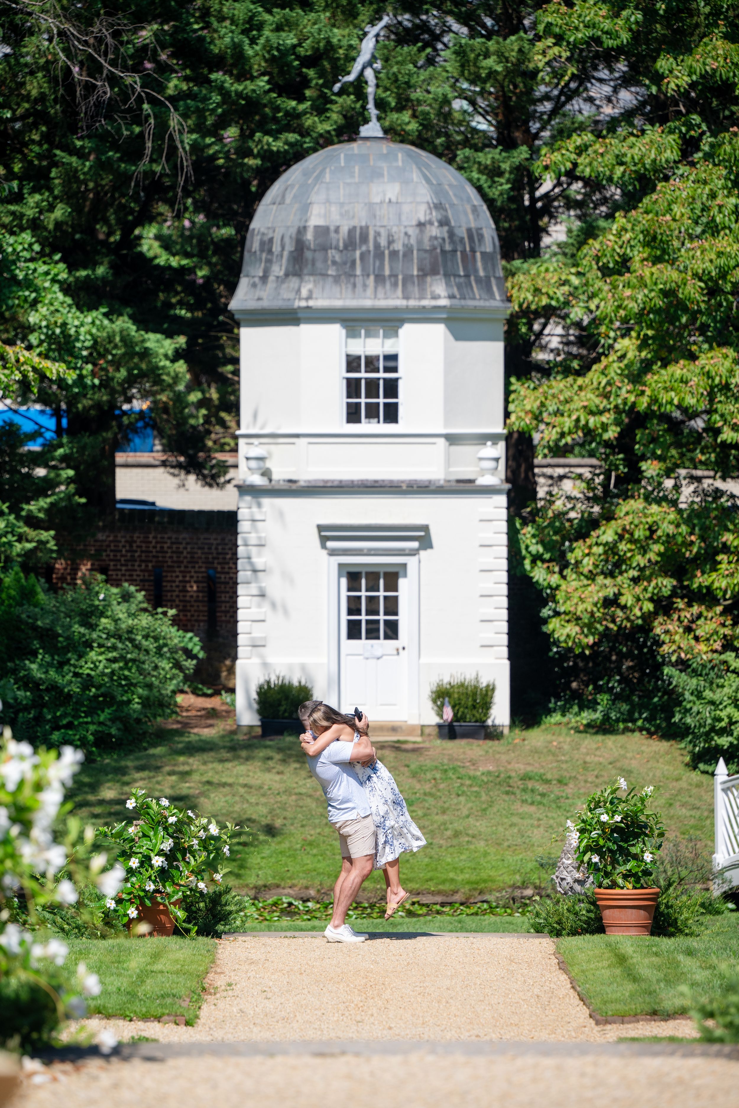Couple embracing on garden path before white domed pavilion surrounded by lush green trees and manicured lawn.