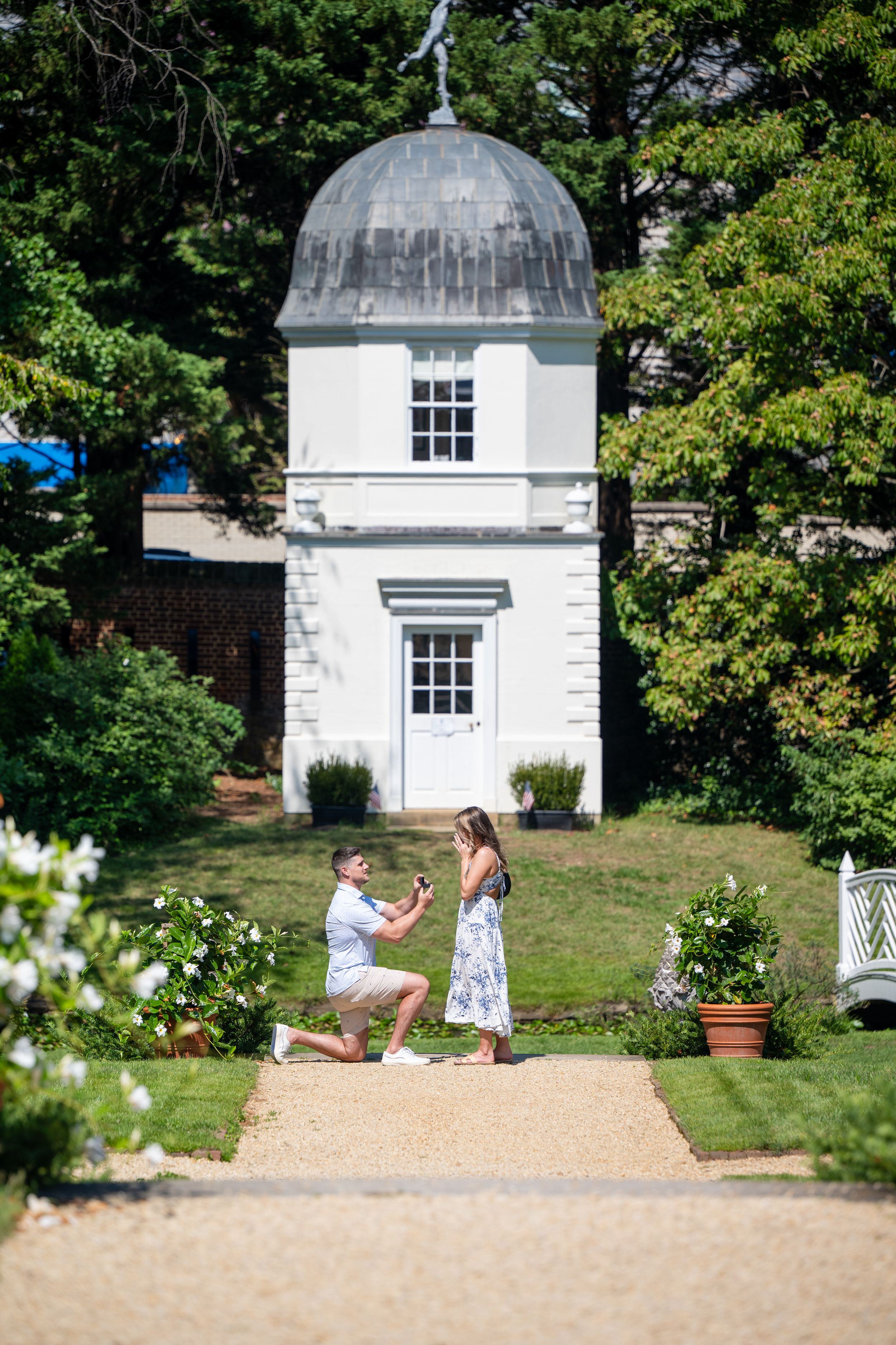 Man proposes on one knee to woman on garden path before white domed gazebo on sunny day.