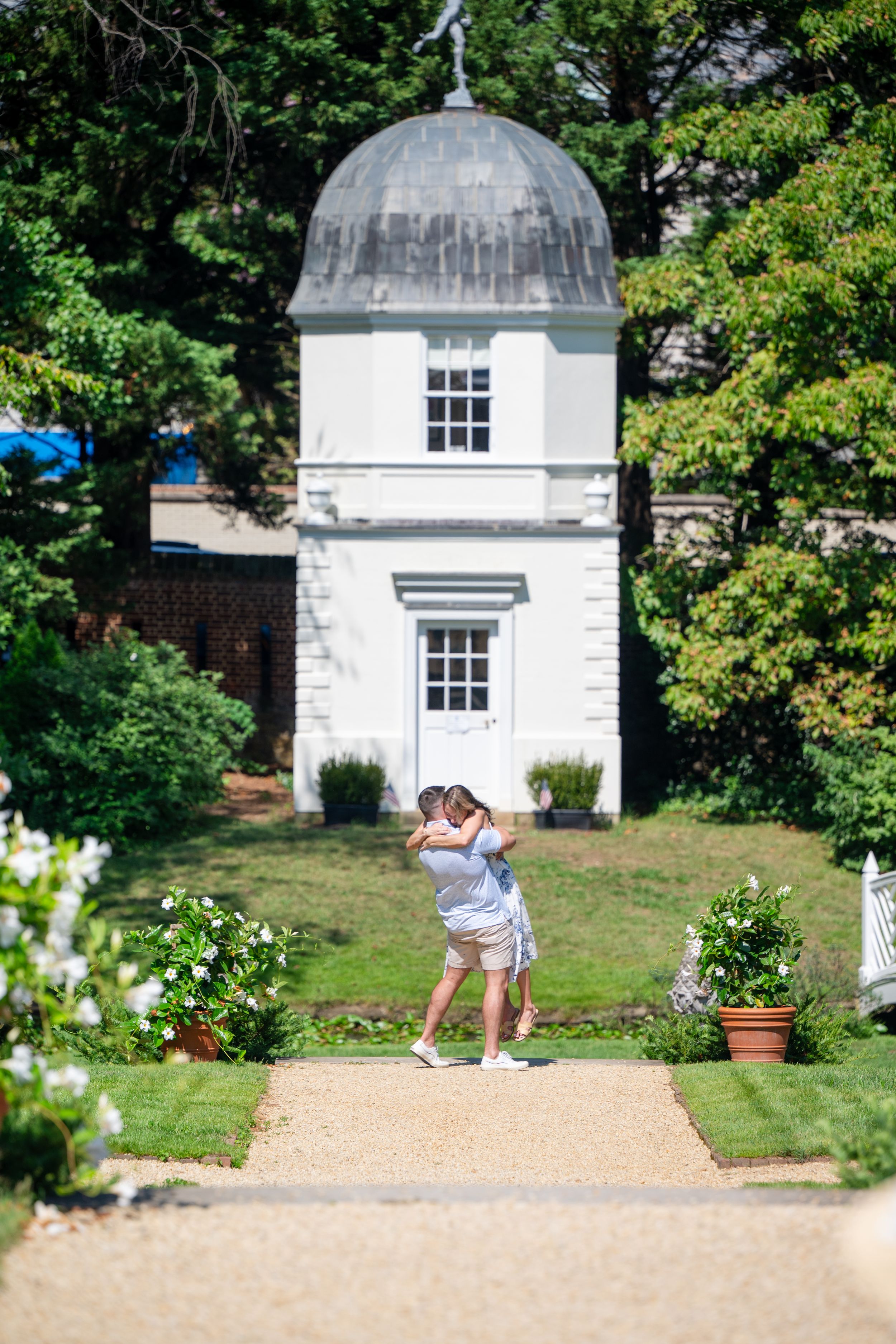 Couple embracing in a formal garden with a white gazebo and lush greenery in the background.