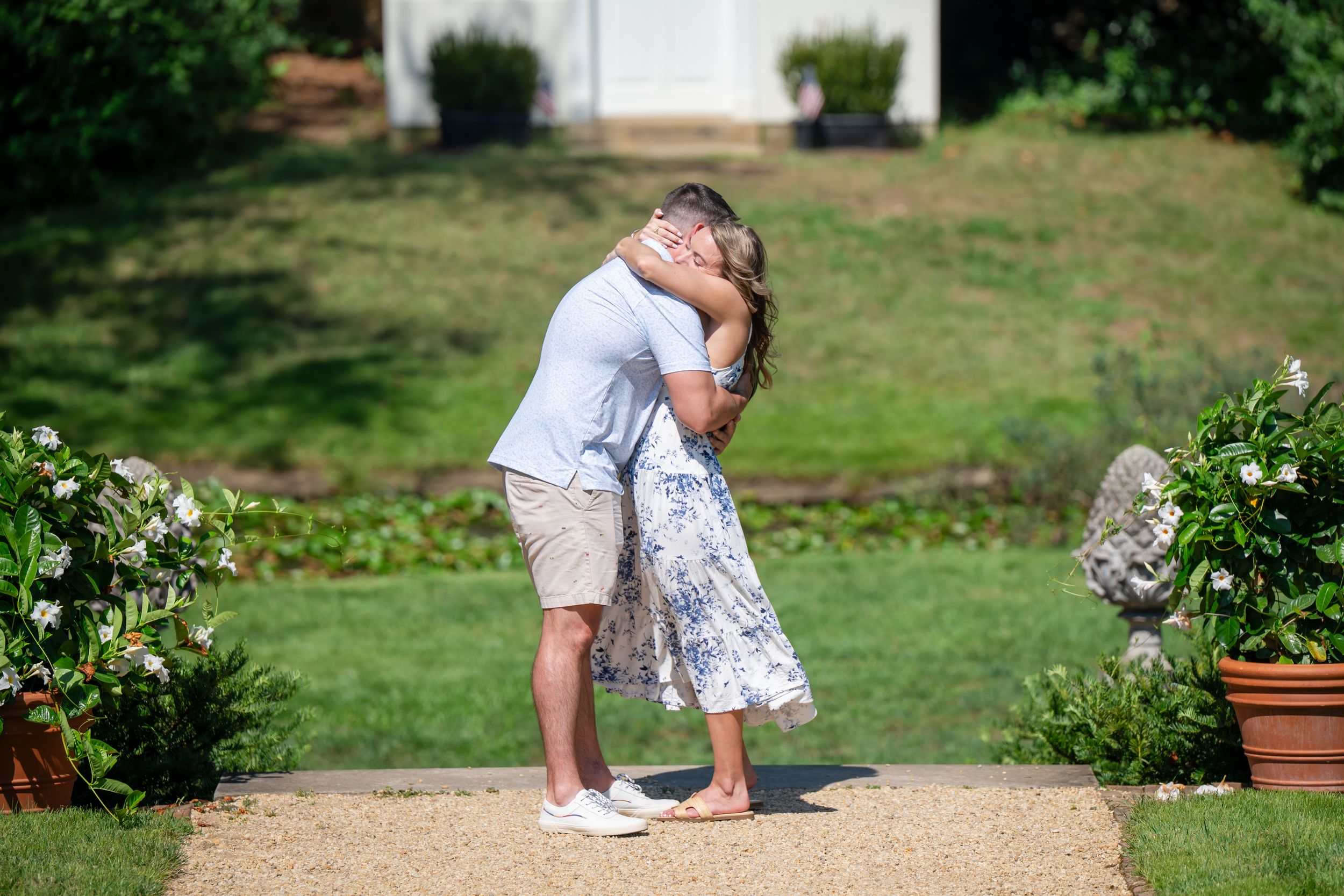 Couple sharing a romantic kiss on a garden pathway surrounded by lush greenery and flowers on a sunny day.