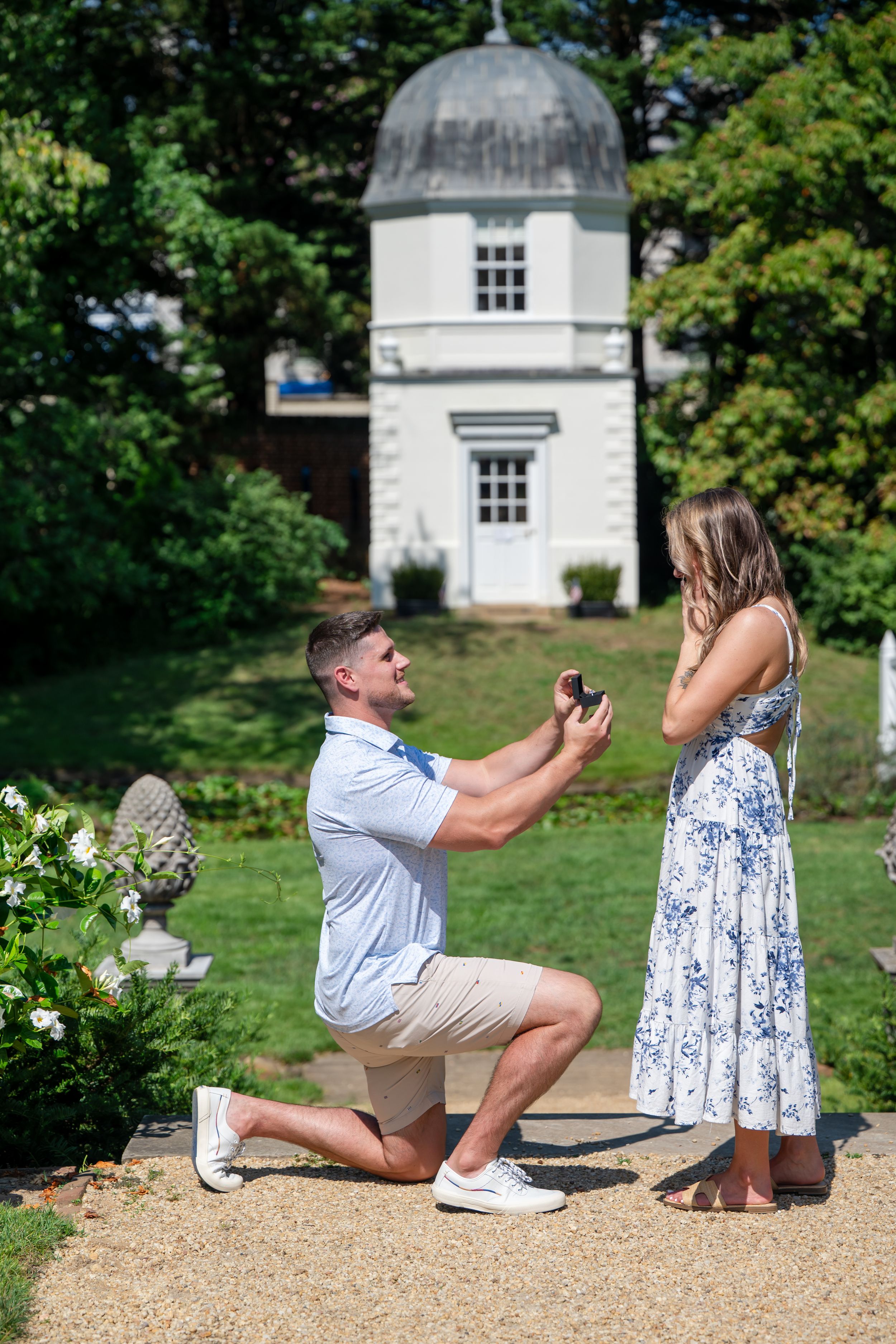 Man proposes on one knee to woman in floral dress in sunny garden near white gazebo.