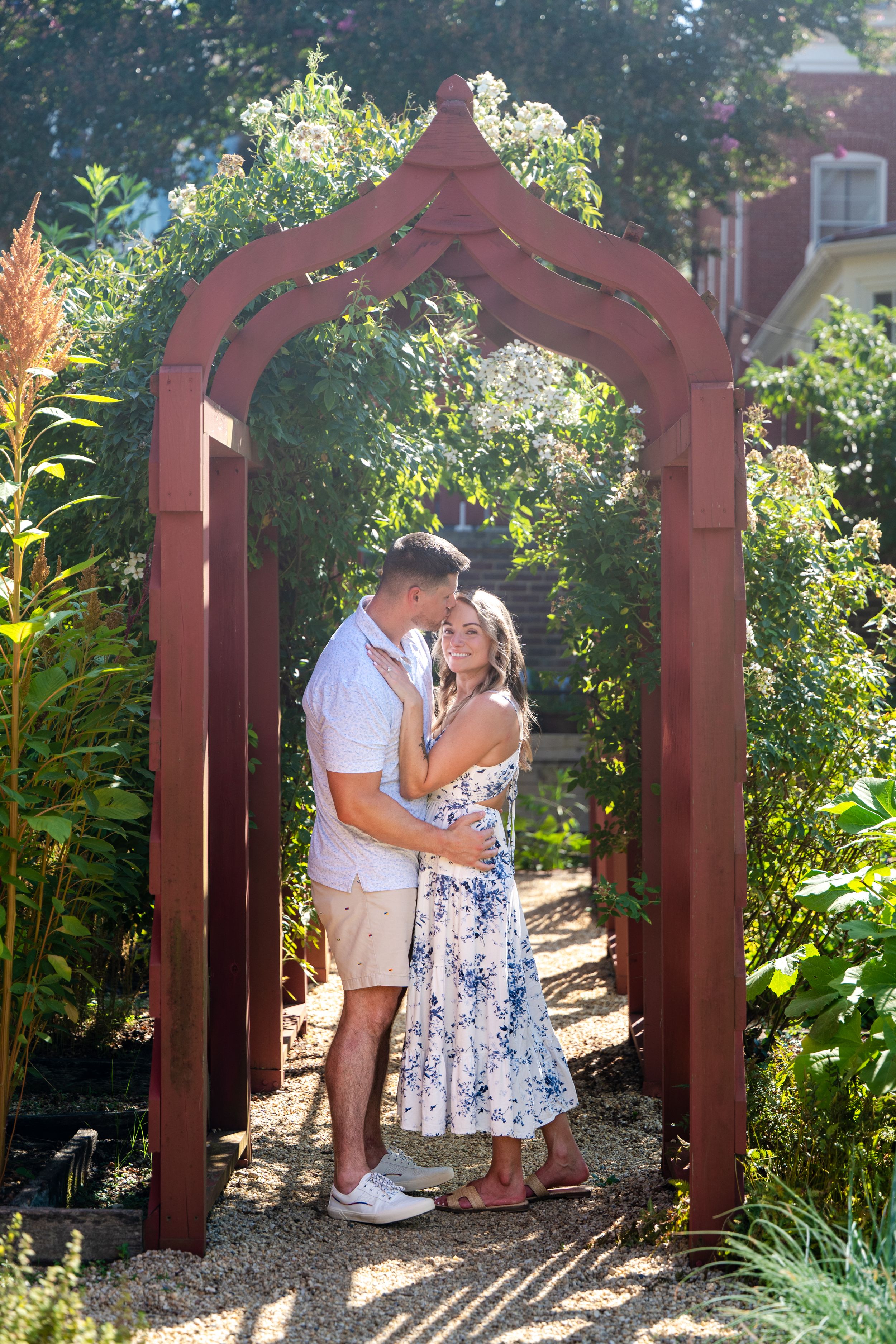 Couple embracing under a red wooden garden arch surrounded by lush greenery on a sunny day.
