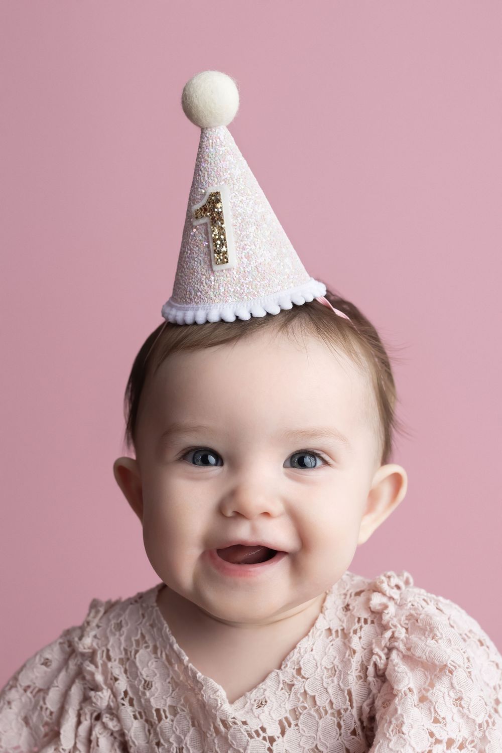 Photo of baby girl wearing a pink birthday hat, smiling, on a pink background, taken by Jen Knightstep, Photographer