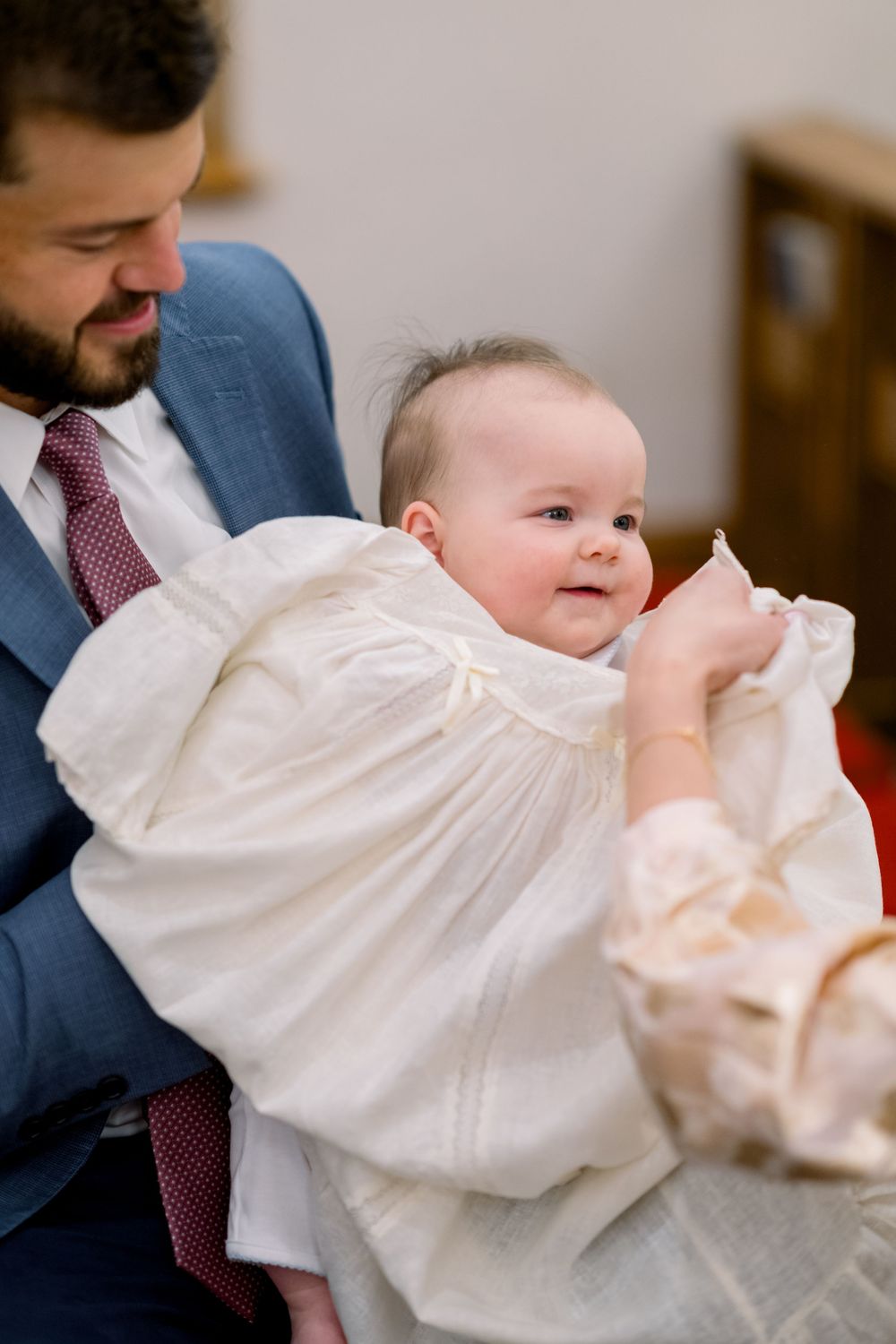 A baby in a flowing white christening gown smiles while being held in formal attire at a baptism ceremony.