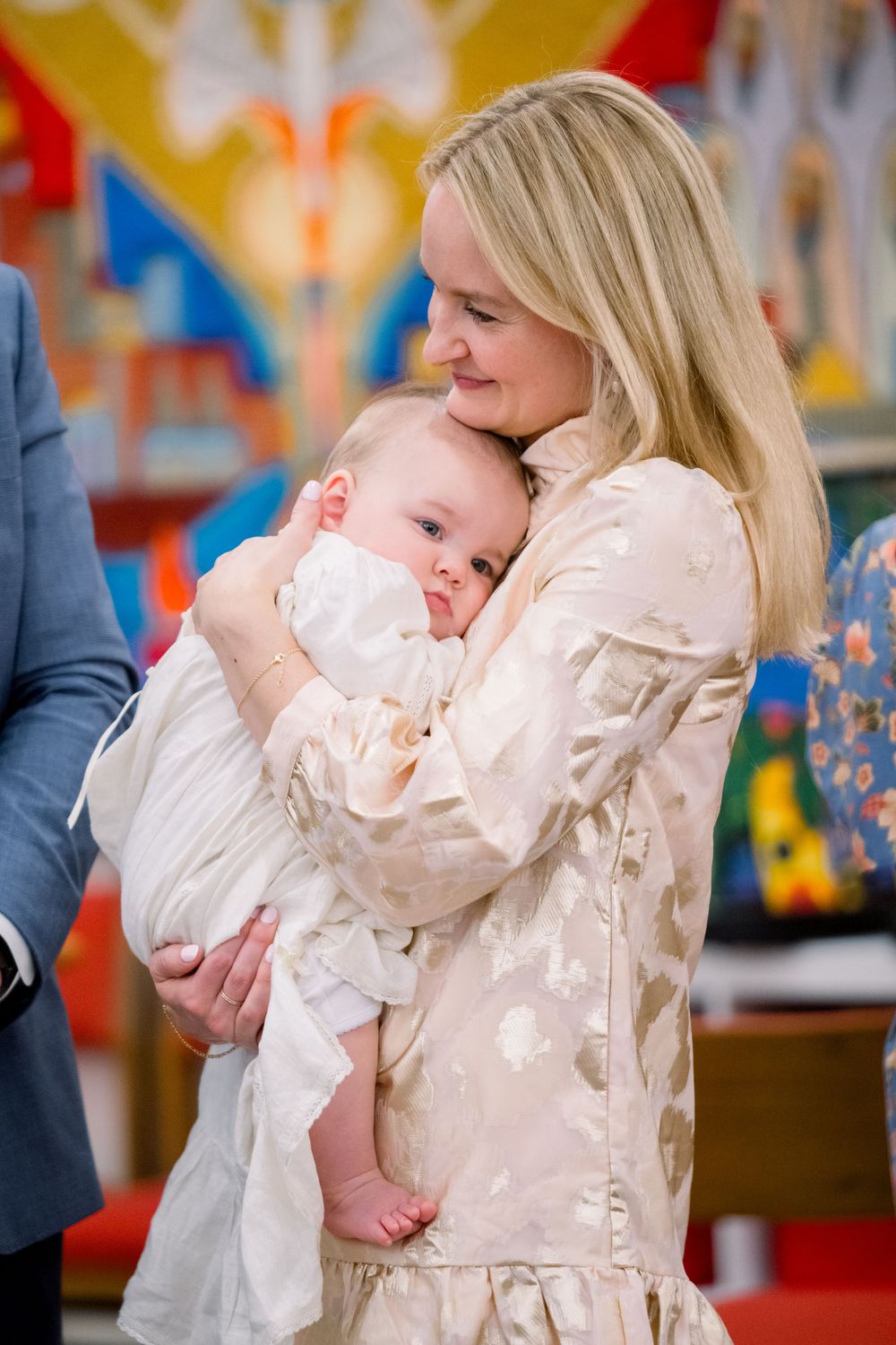 A tender moment captured during a christening ceremony with a baby in white against a colorful background.