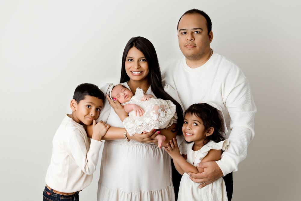 Family of five newborn session portrait in white and cream neutral tones. Newborn baby sleeping in mothers arms with siblings and dad around