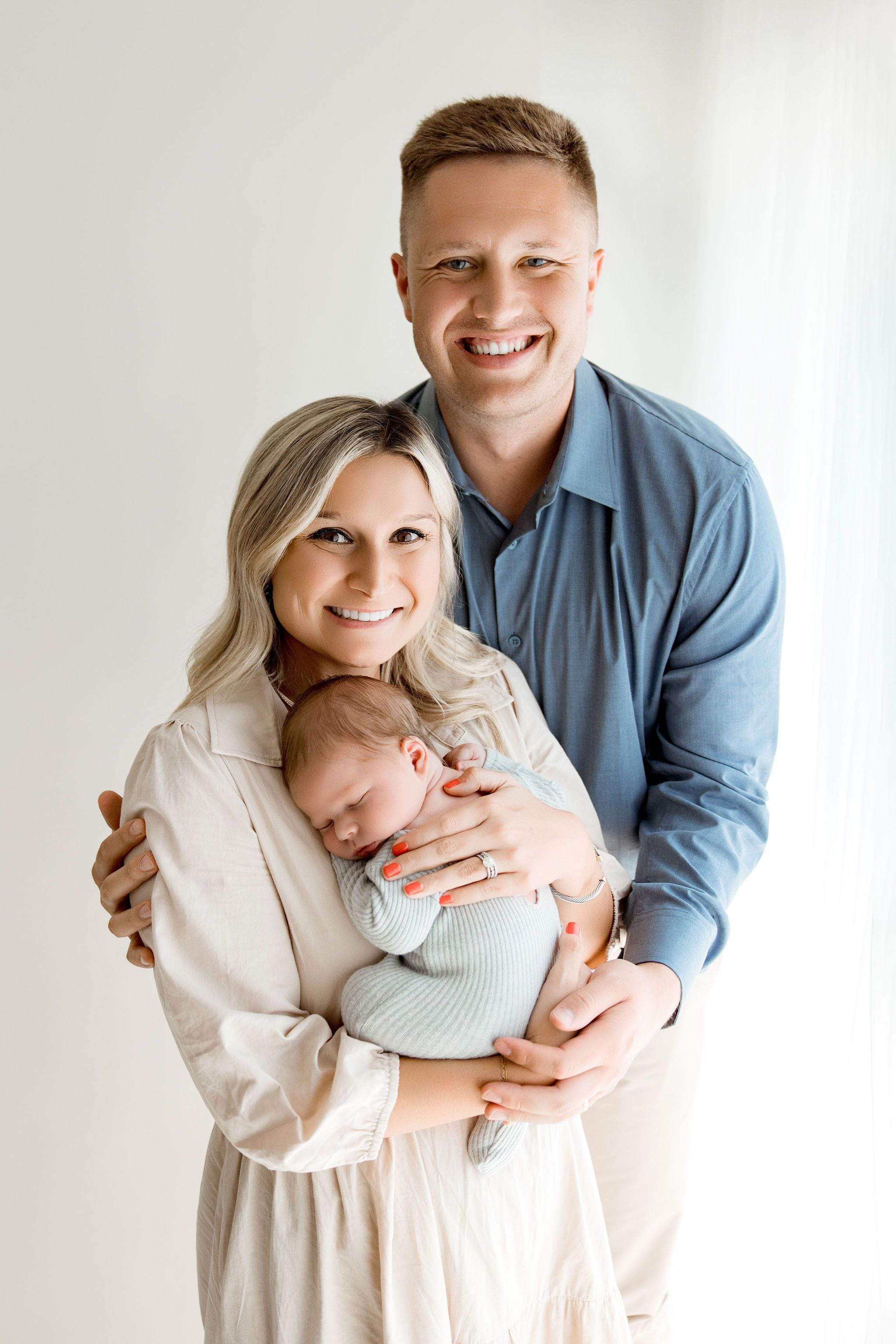 Family of three posed by the window with baby on in mom's arms. Schaumburg IL Newborn Session