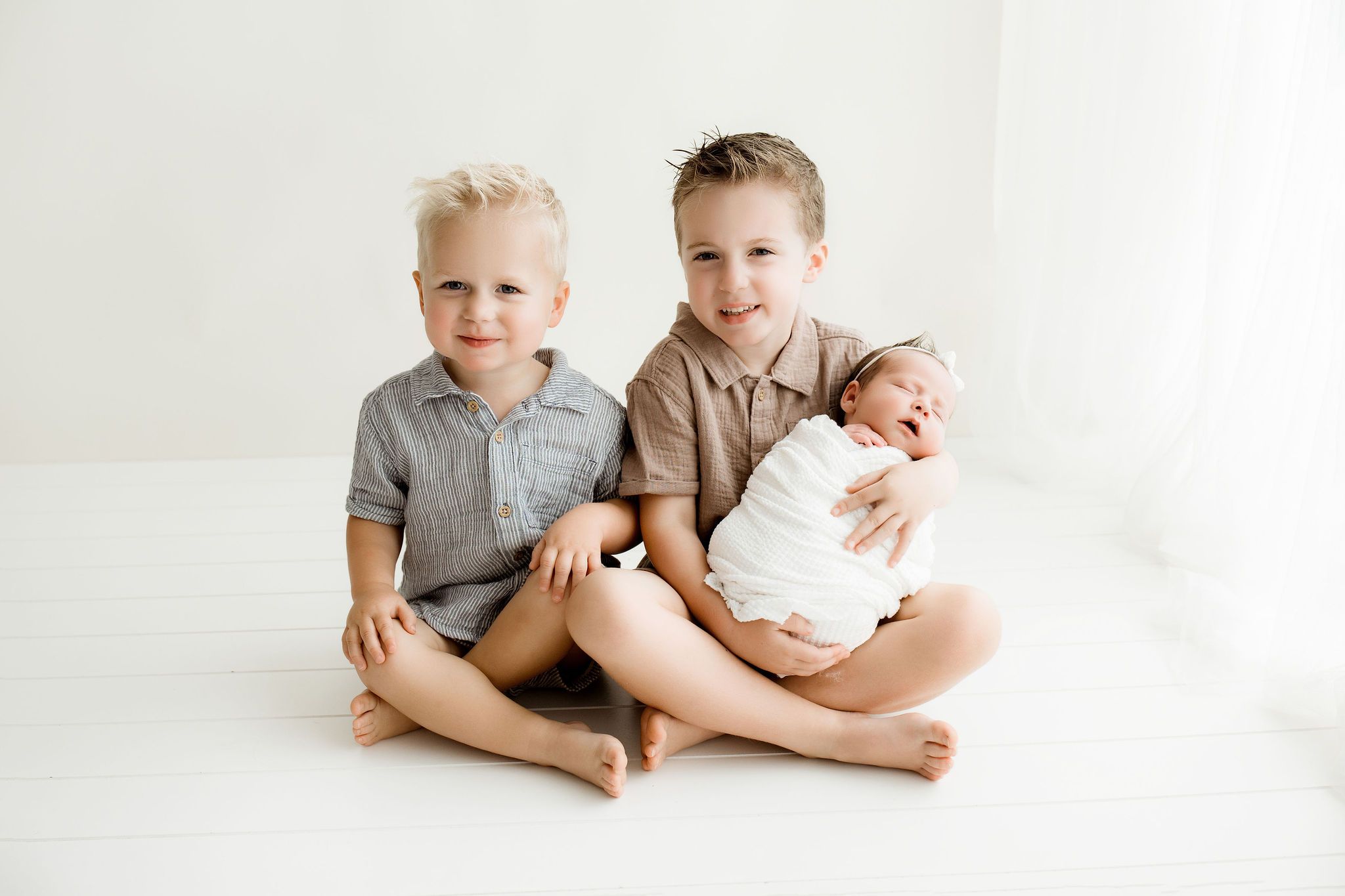 Two boys, older brothers holding their newborn sister while sitting by the window. Newborn session