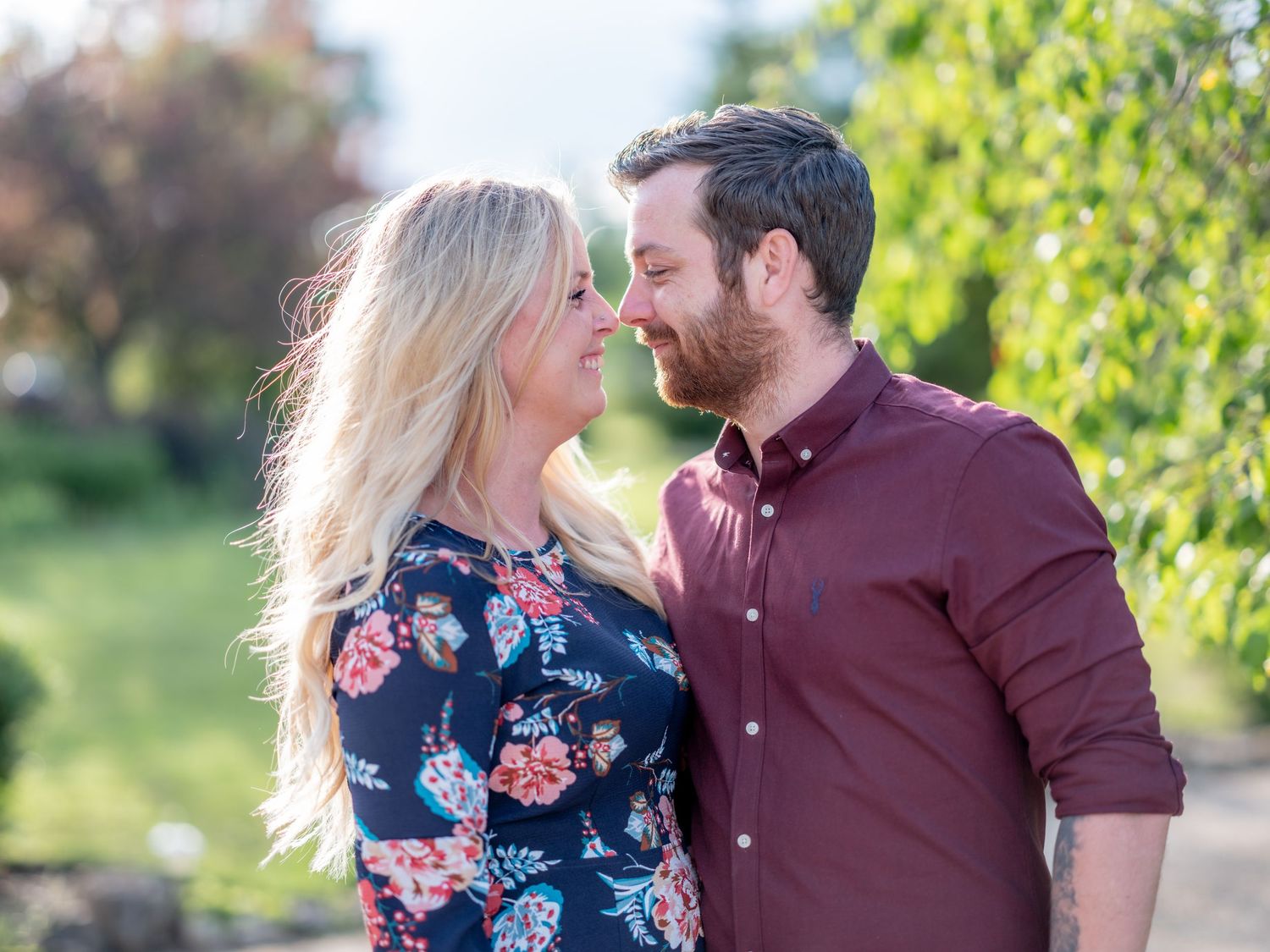 A couple in romantic poses outdoors, with the woman wearing a navy floral dress and the man in a burgundy shirt.