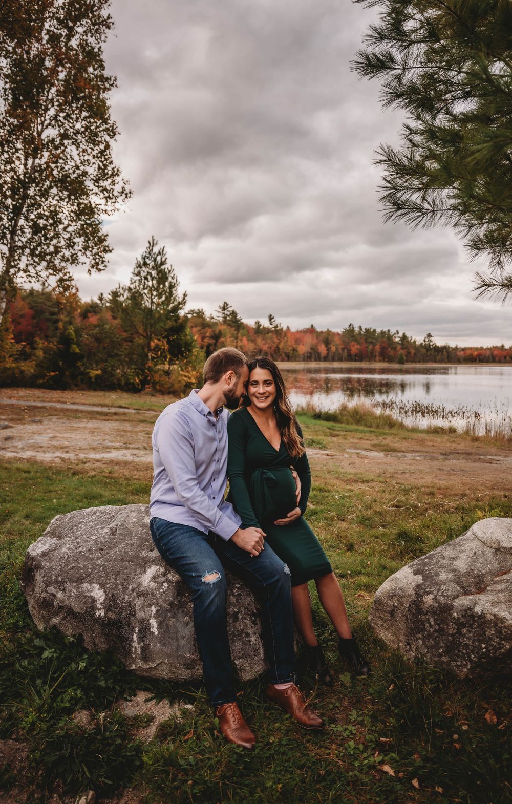 A couple sits on large rocks by an autumn lakeside with pine trees and colorful fall foliage in the background.