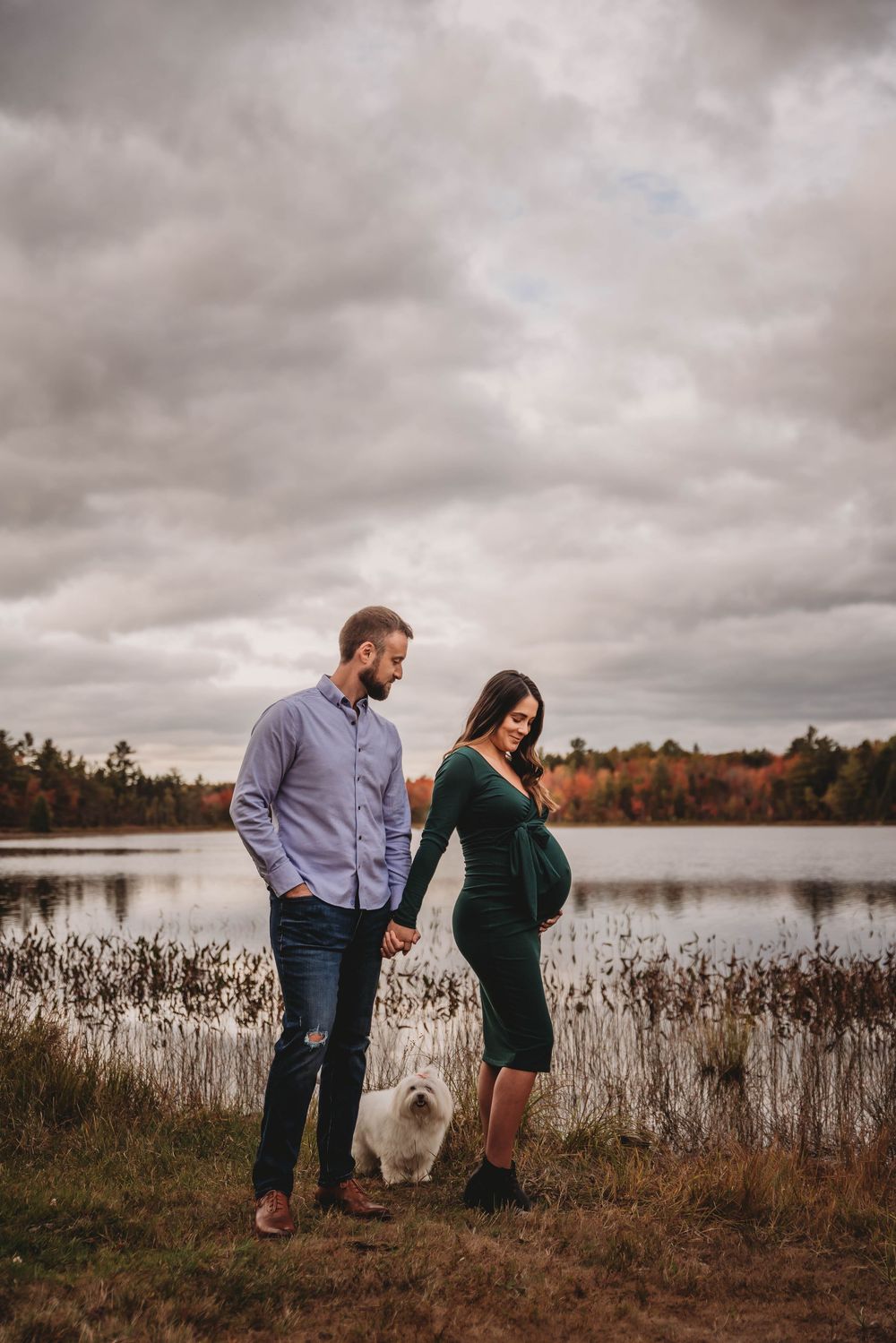 A couple and their small white dog stand by an autumn lake under dramatic cloudy skies.