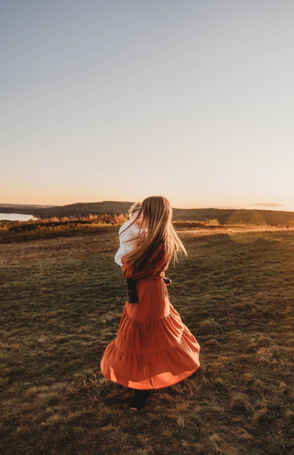 A figure in a flowing orange maxi dress twirls on a hilltop against a golden sunset landscape.