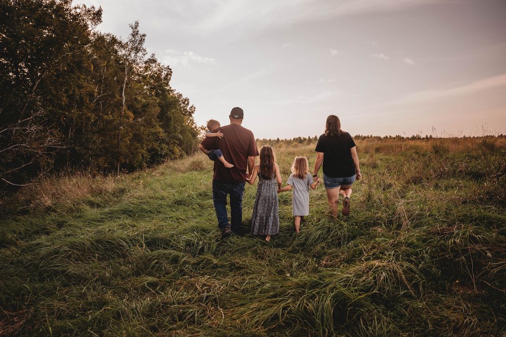 A family walks together through a grassy meadow on a late summer evening with trees in the background.