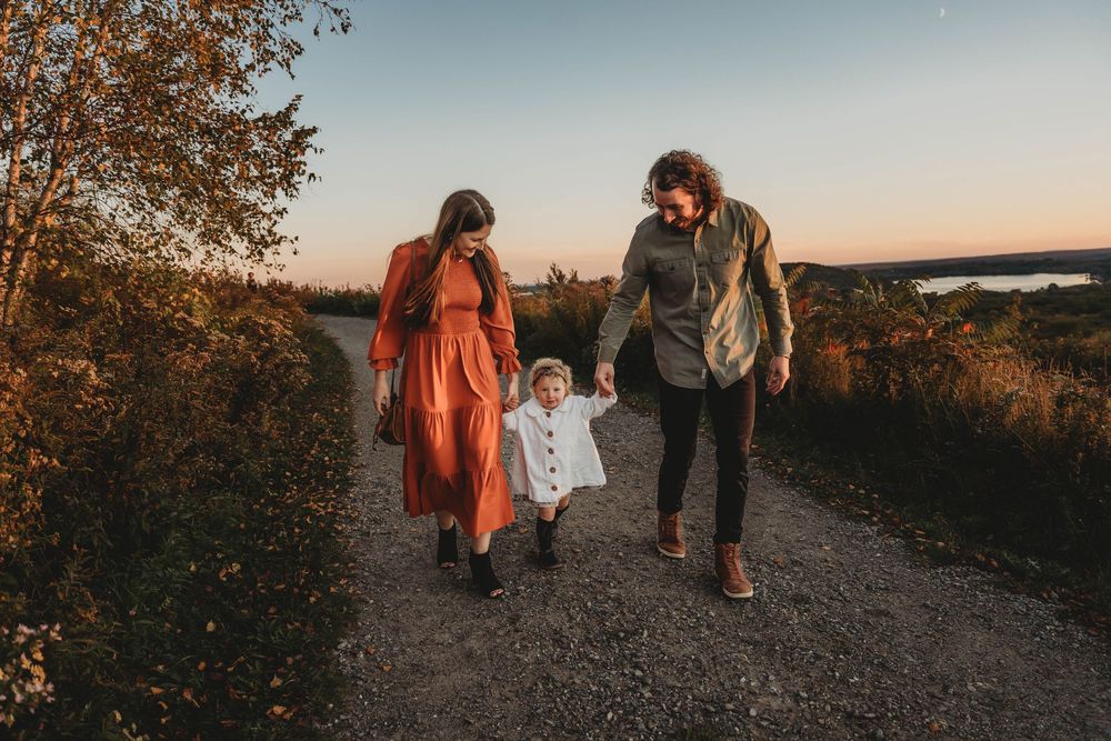 A family walks together on a gravel path during sunset, with autumn colors and water in the background.
