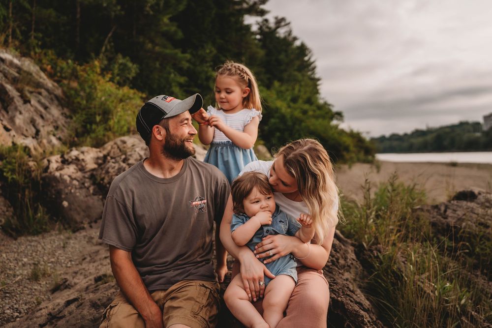 A loving family sits together on rocky beach terrain at sunset with trees and cloudy skies in the background.