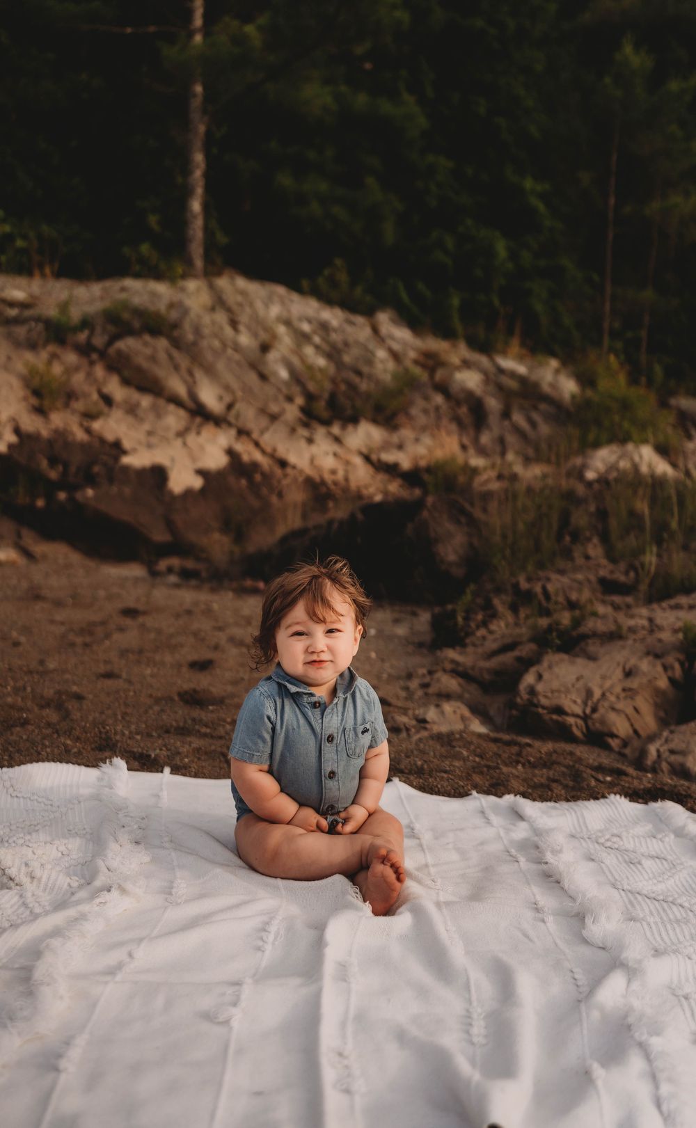 Small child sits on a white blanket outdoors against a rocky natural backdrop during golden hour.