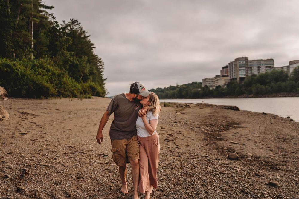 A couple walks intimately along a sandy beach shoreline with trees and buildings in the distance on an overcast day.