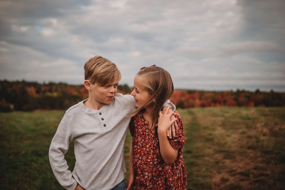 Two young people share a tender moment together outdoors with autumn trees and cloudy sky in the background.