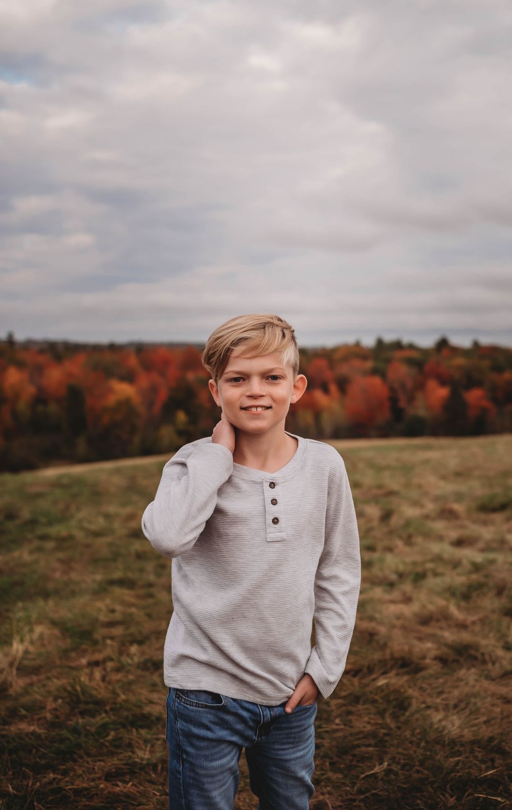A young person in a grey henley sweater stands in a field with vibrant autumn foliage in the background.