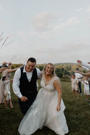 Wedding couple laughing joyfully on hillside while guests wave in background.