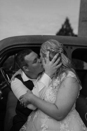 Black and white photo of intimate moment between newlyweds near vintage car.