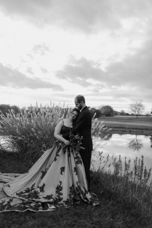 A black and white portrait of a couple in formal wear by a pond with tall grasses.