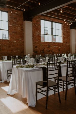 An industrial wedding reception venue with exposed brick walls, white tablecloths and black chiavari chairs.