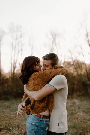 A romantic embrace outdoors with one person wearing a cozy brown sweater and blue jeans against an autumn backdrop.