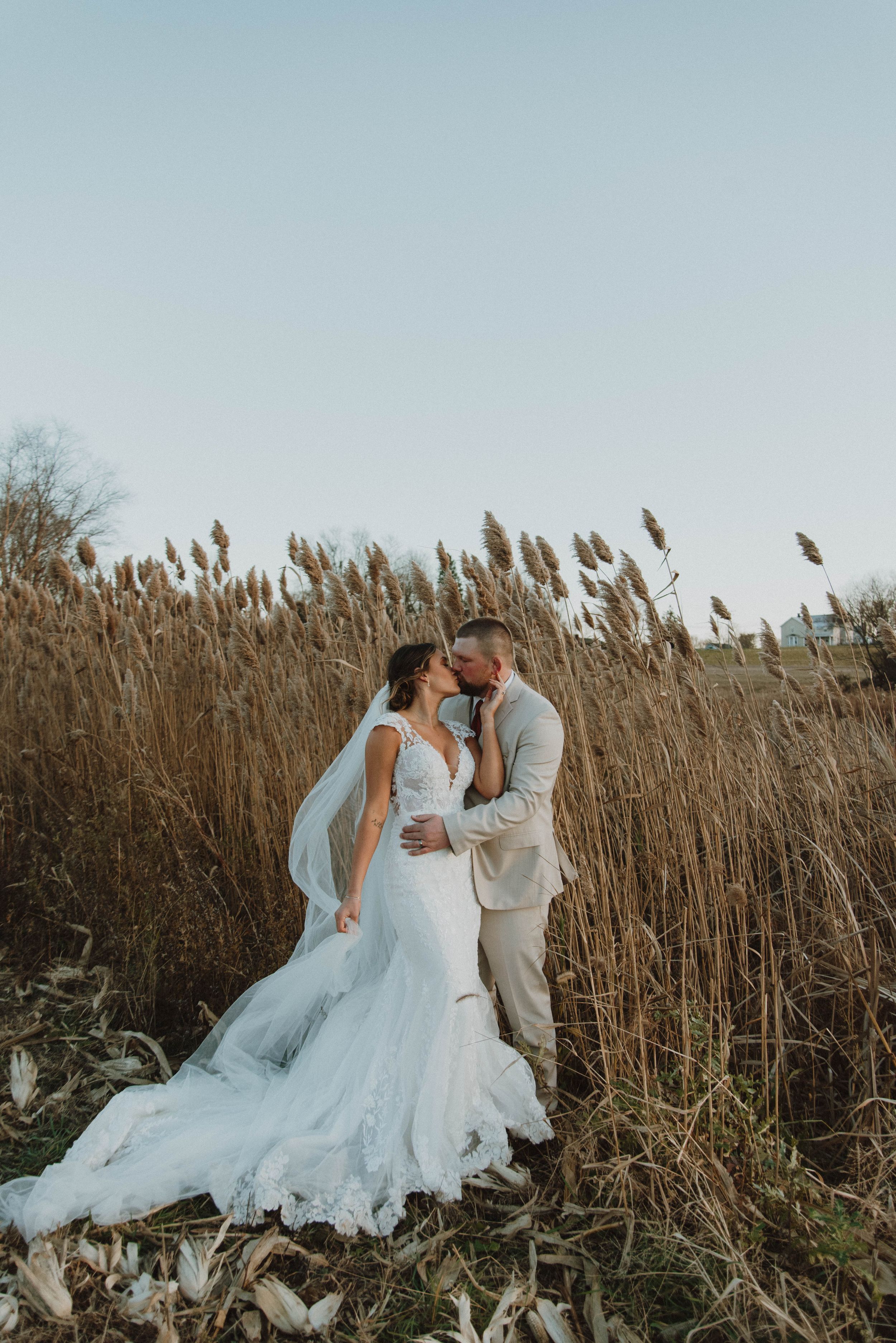 A couple embraces among tall grass with a flowing white wedding dress.