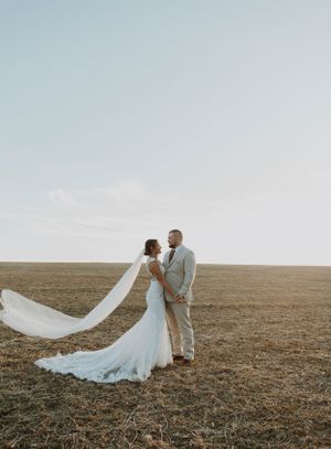 Wedding couple embraces in an open field with dramatic flowing veil catching the sunlight.