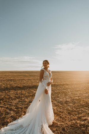 Bride in flowing lace gown poses alone in golden sunset light on open field.
