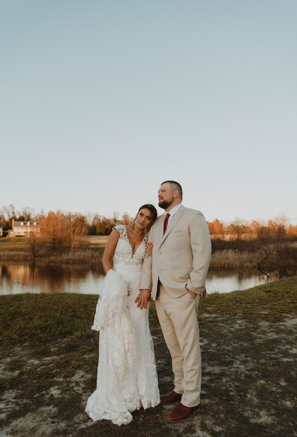 Wedding couple in elegant attire poses at sunset by a lake with autumn foliage in the background.