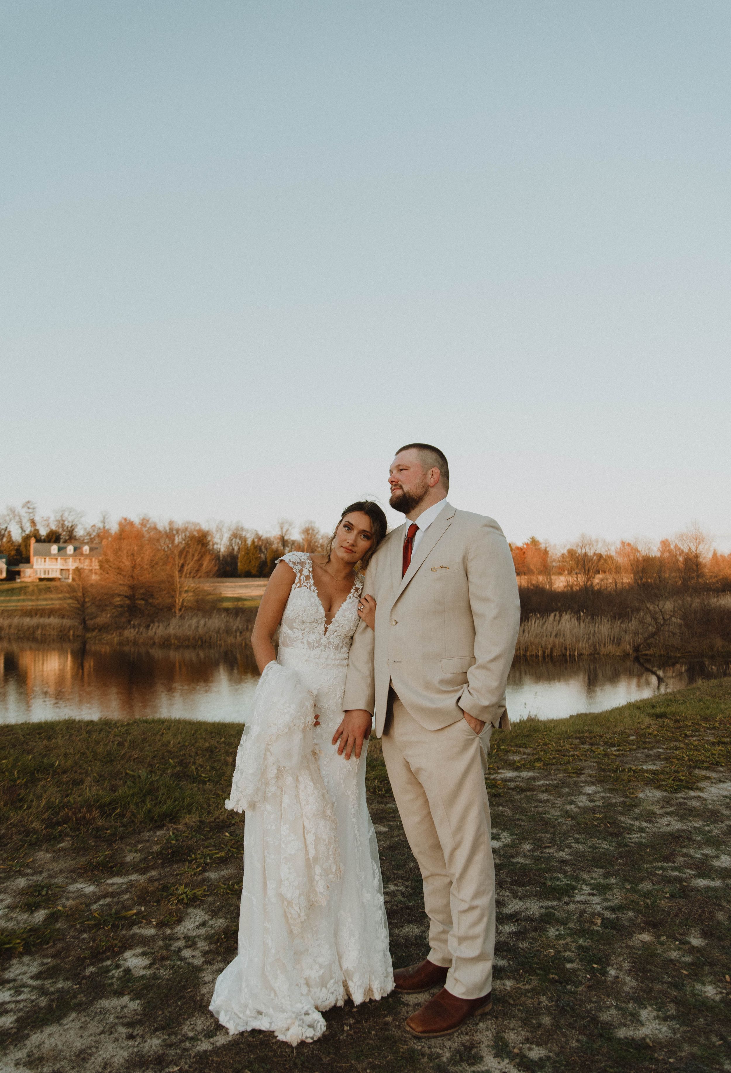 Wedding couple in elegant attire poses at sunset by a lake with autumn foliage in the background.