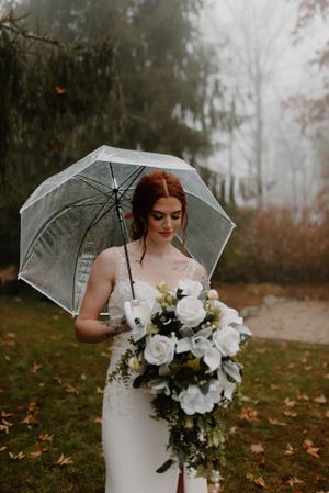 Bride with white bouquet poses with clear umbrella in misty autumn setting.