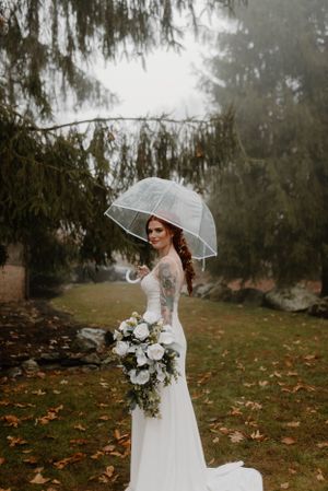 A bride in a long-sleeved white dress holds a white and green bouquet while posing with a clear umbrella on a misty day.